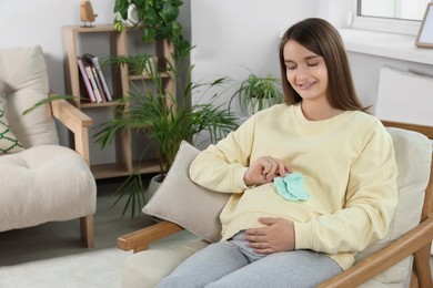 Happy pregnant woman sitting on armchair with baby socks in living room