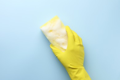 Cleaner in rubber glove holding sponge with foam on light blue background, top view