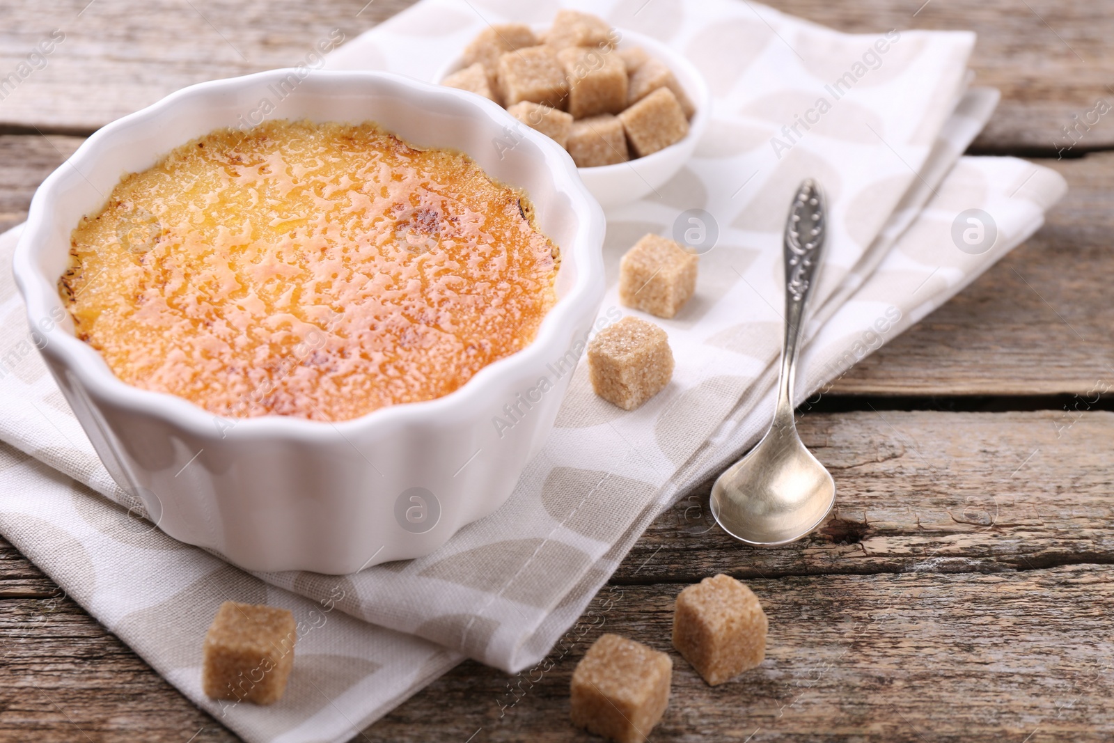 Photo of Delicious creme brulee in bowl, sugar cubes and spoon on wooden table, closeup