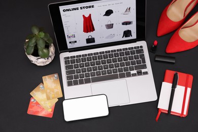 Photo of Online store website on laptop screen. Computer, smartphone, credit cards, women's shoes, stationery and lipstick on black background, above view