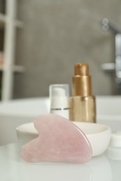 Photo of Rose quartz gua sha tool and cosmetic products on white table in bathroom