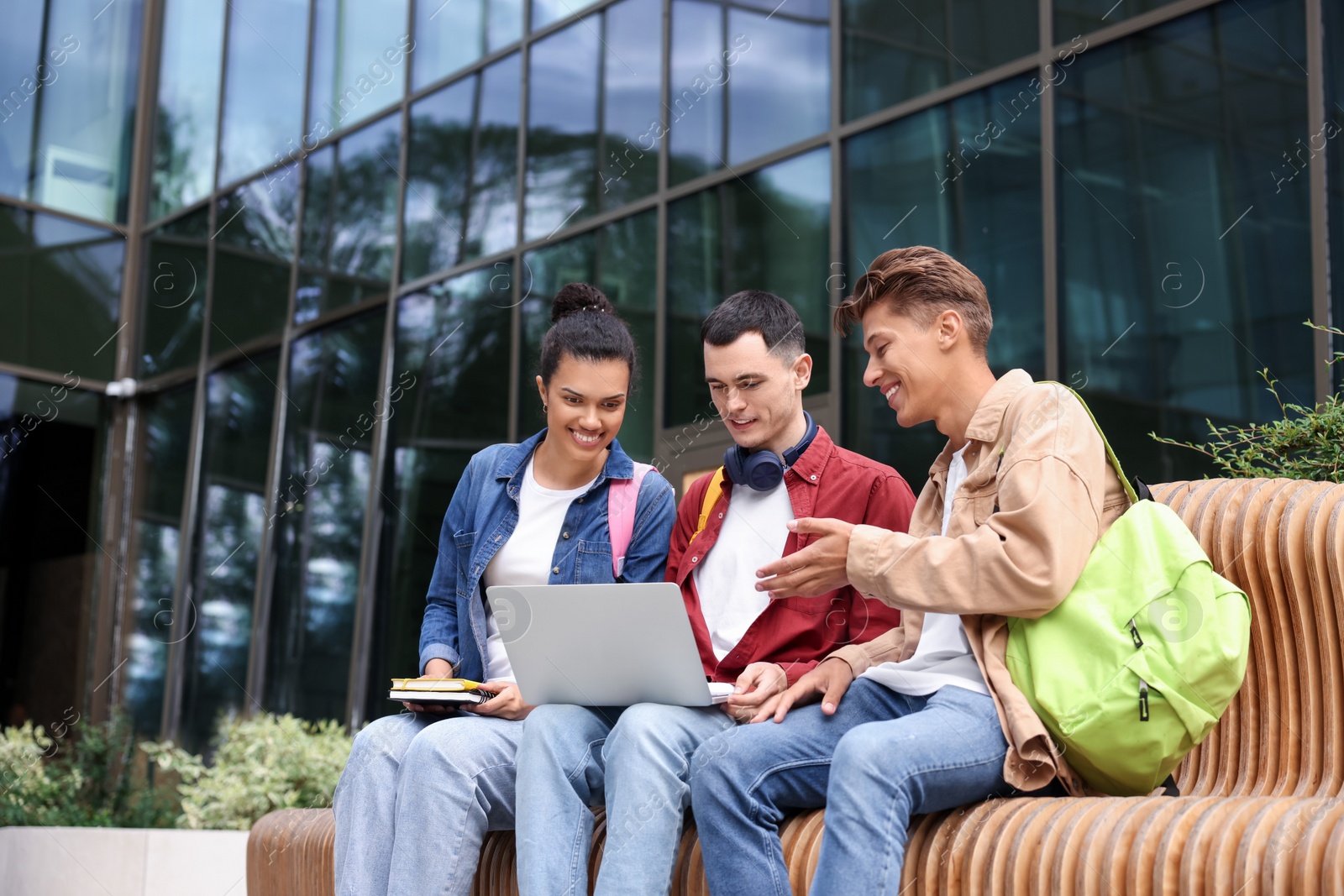 Photo of Happy young students studying with laptop on bench outdoors