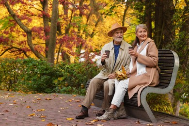 Affectionate senior couple with cups of coffee spending time together on bench in autumn park, space for text