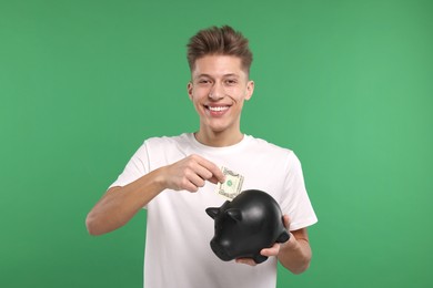 Photo of Happy man putting dollar banknote into piggy bank on green background