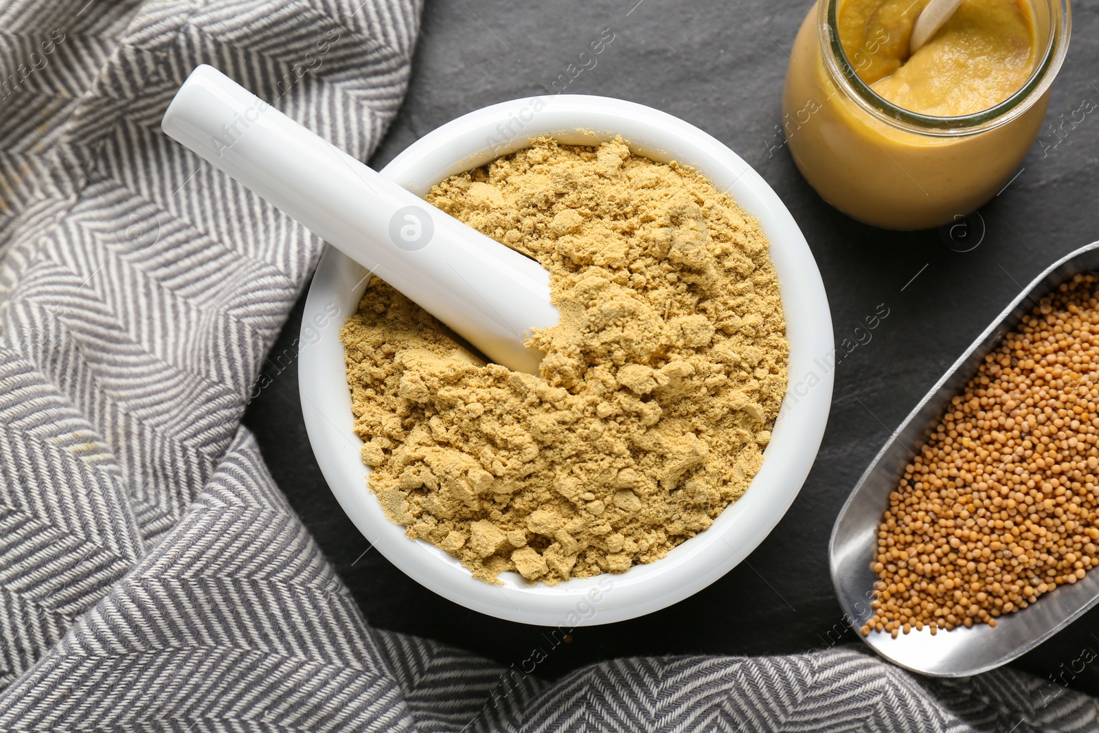 Photo of Mortar of mustard powder and scoop with seeds on black table, flat lay