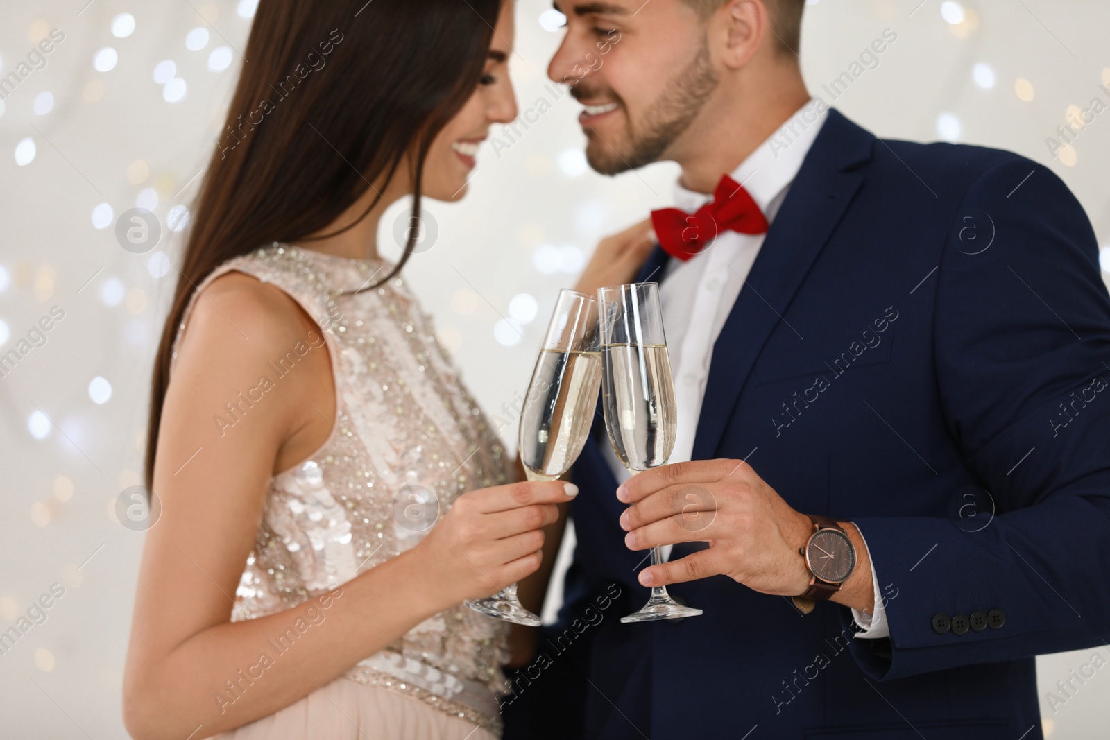 Photo of Lovely young couple with glasses of champagne against blurred festive lights, closeup. Christmas celebration