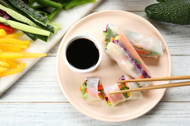 Photo of Delicious rolls wrapped in rice paper served on white wooden table, flat lay