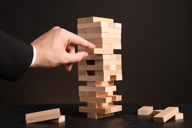 Photo of Playing Jenga. Man removing wooden block from tower at black table against dark background, closeup