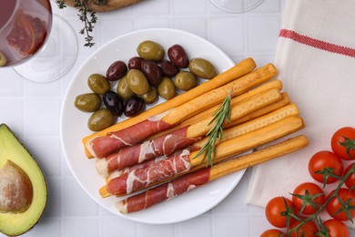 Photo of Delicious grissini sticks with prosciutto and ingredients on white table, flat lay
