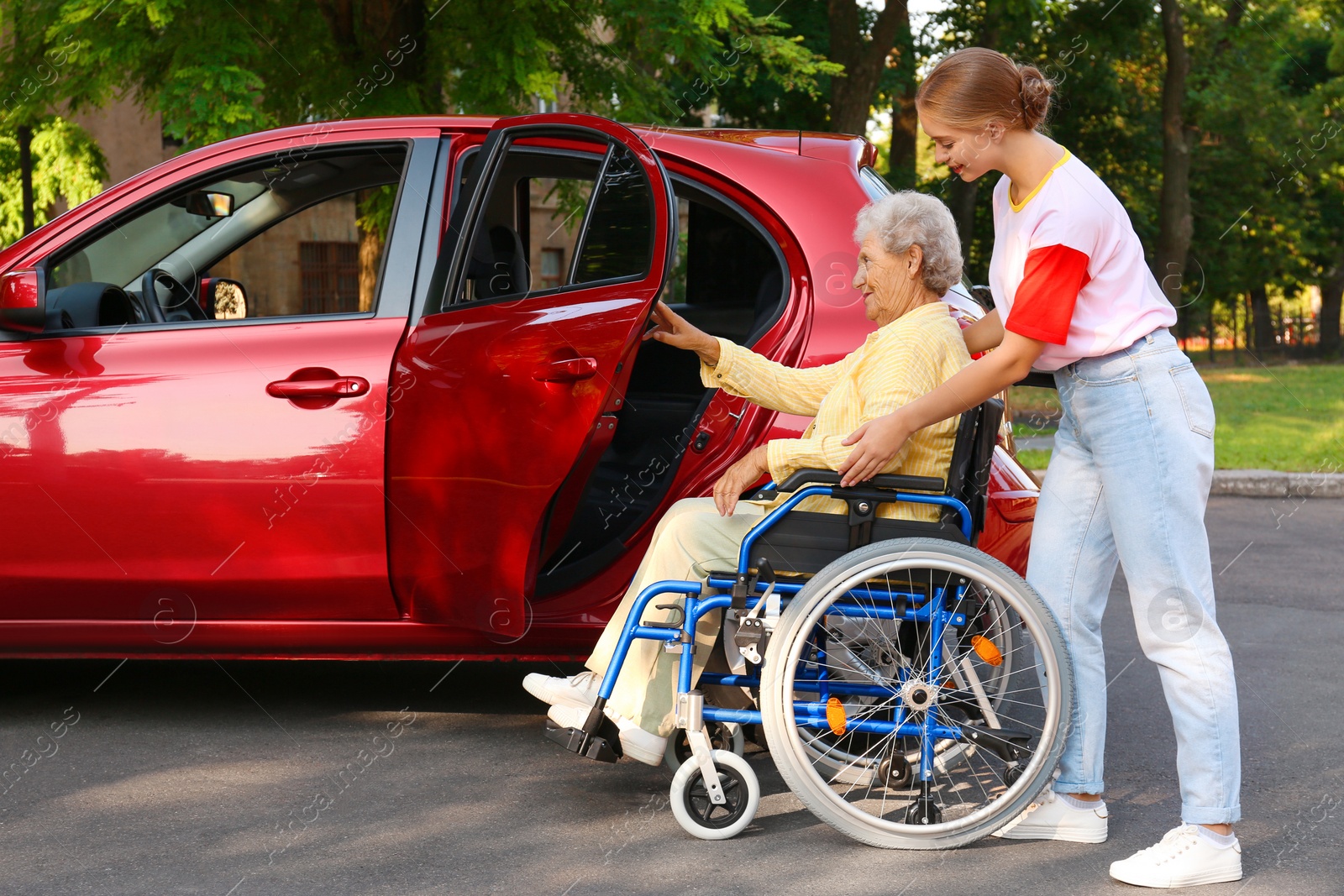 Photo of Young woman helping disabled grandmother in wheelchair to get into car outdoors