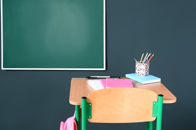 Wooden school desk with stationery and backpack near blackboard on grey wall