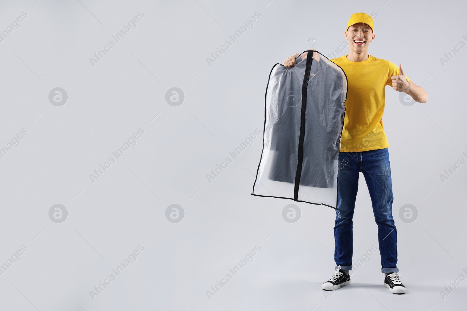 Photo of Dry-cleaning delivery. Happy courier holding garment cover with clothes and showing thumbs up on light grey background, space for text