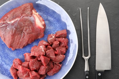 Plate with pieces of raw beef meat, knife and fork on grey textured table, flat lay