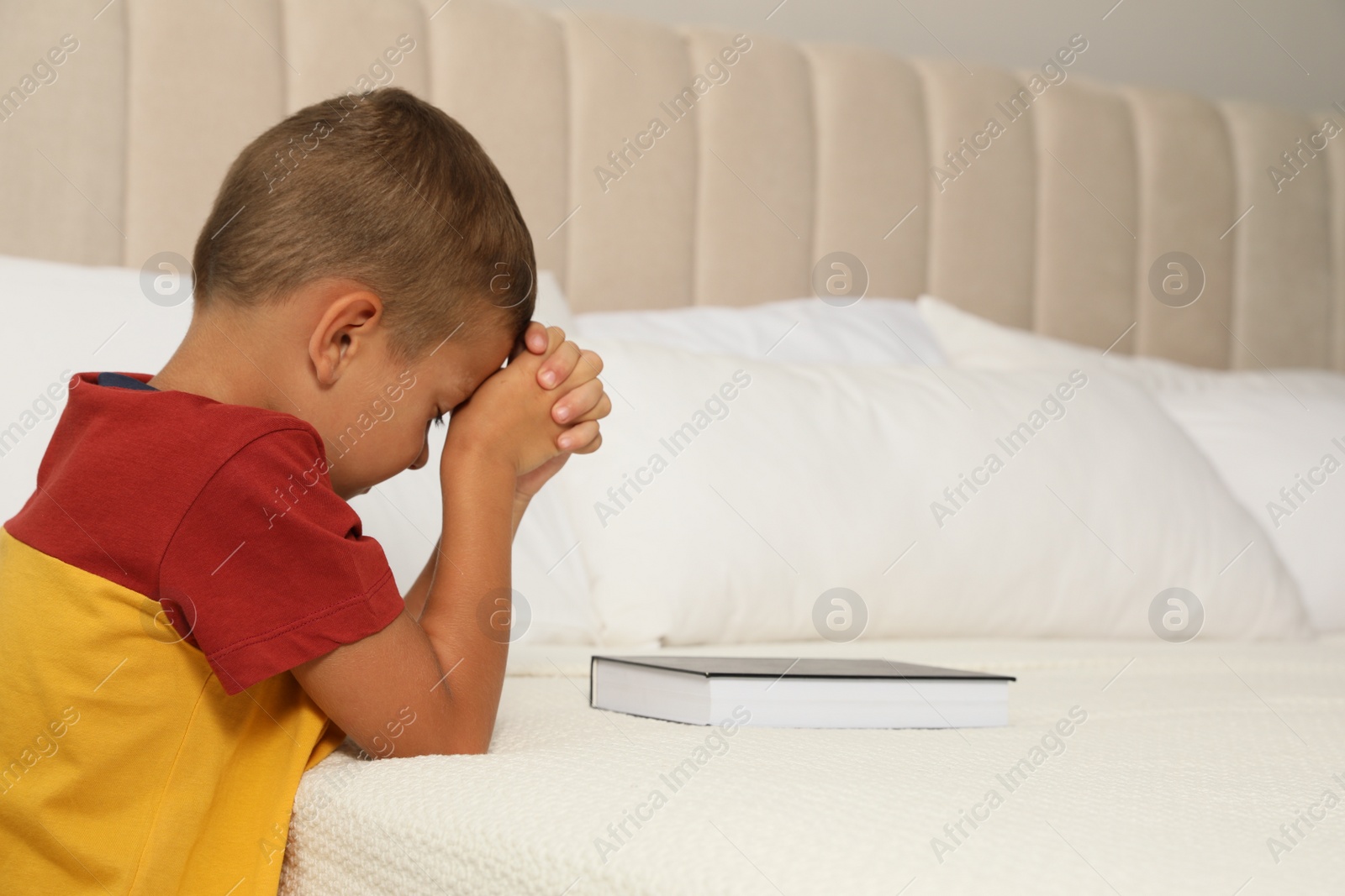 Photo of Cute little boy with hands clasped together saying bedtime prayer over Bible at home. Space for text