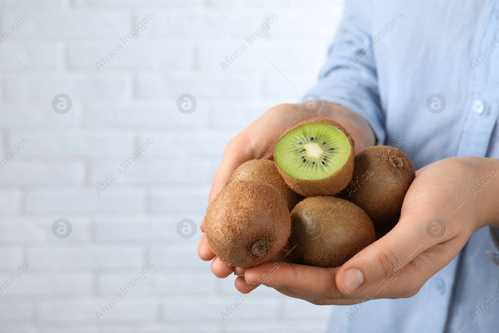 Photo of Woman holding pile of fresh ripe kiwis against white brick wall, closeup. Space for text