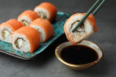 Dipping tasty sushi roll with salmon into soy sauce on grey table, closeup