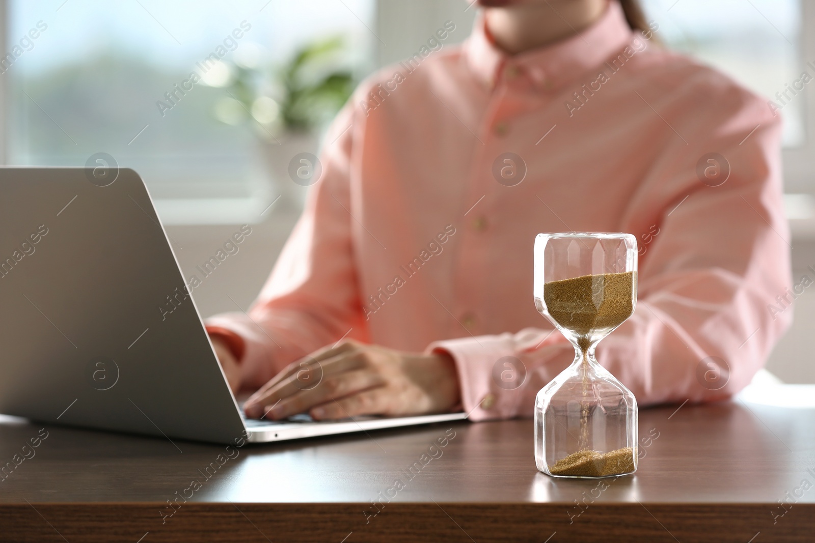 Photo of Hourglass with flowing sand on wooden table, selective focus. Man using laptop indoors