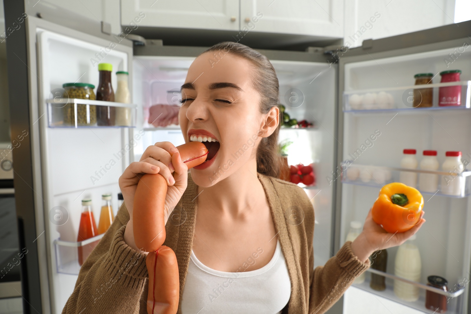 Photo of Young woman eating sausage near open refrigerator