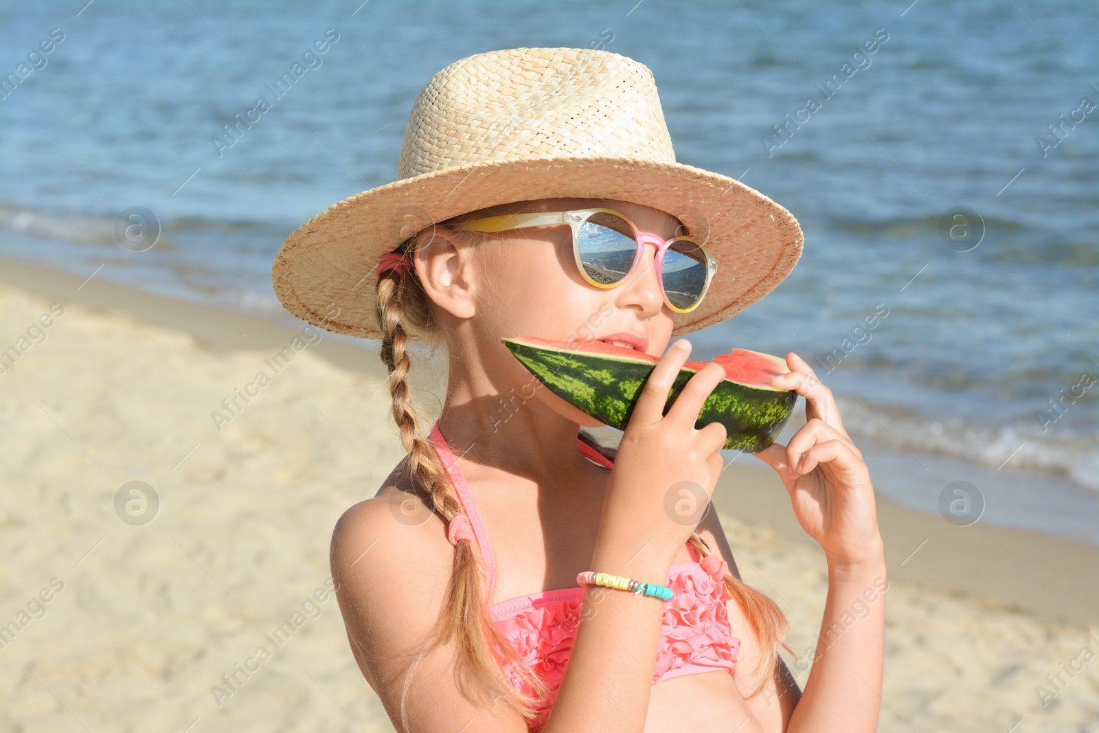 Photo of Cute little girl in straw hat and sunglasses eating juicy watermelon on beach