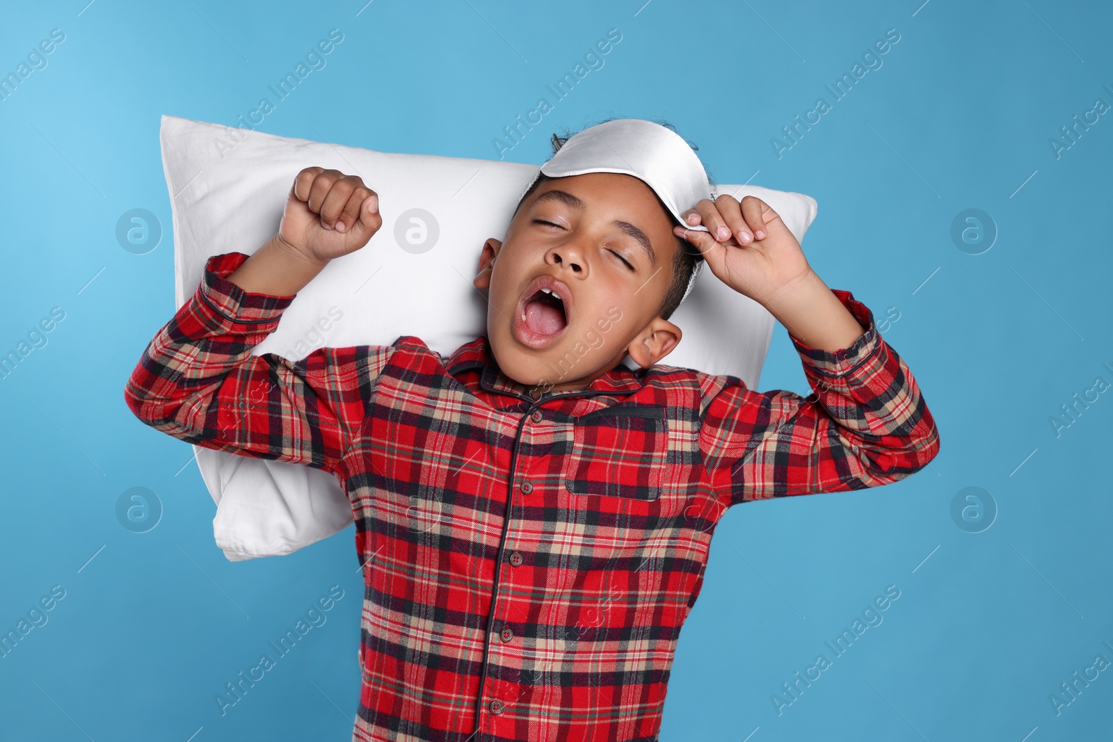 Photo of Boy with pillow and sleep mask yawning and stretching on light blue background. Insomnia problem