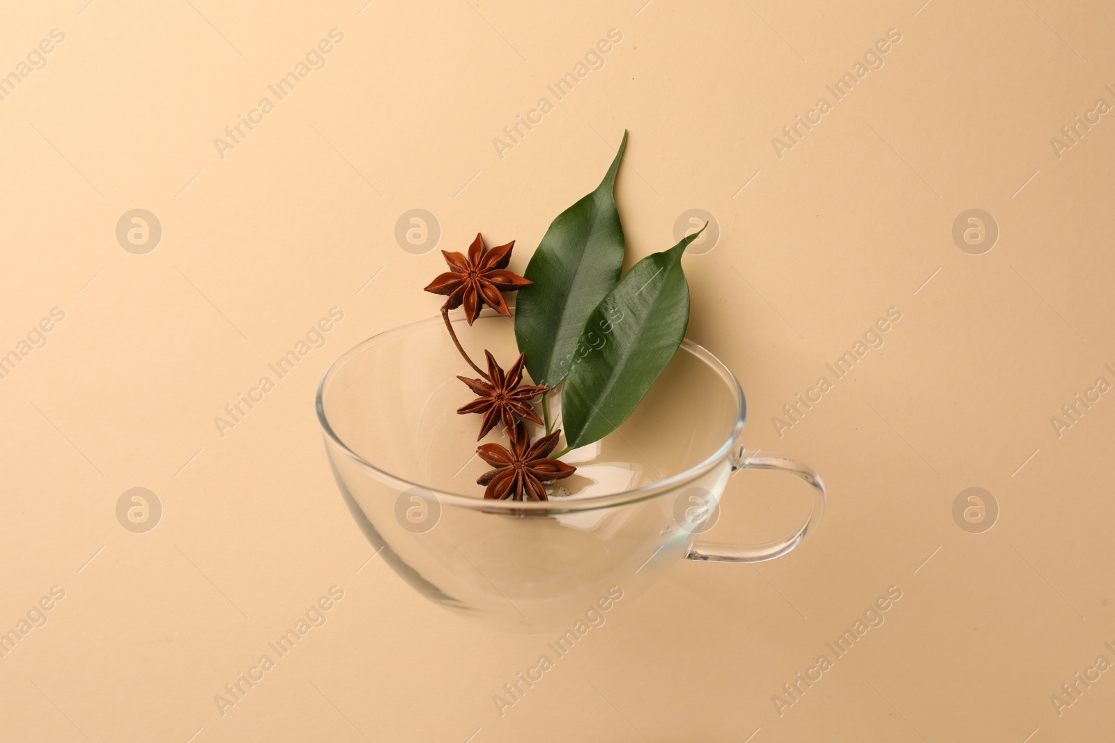 Photo of Anise stars and green leaves falling into glass cup on beige background, flat lay