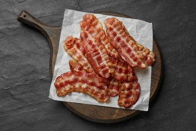 Photo of Board with fried bacon slices on dark textured table, top view
