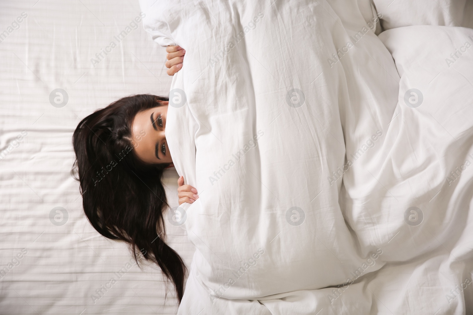 Photo of Young woman lying in bed covered with white blanket, top view