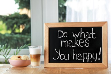 Photo of Blackboard with phrase Do What Makes You Happy, coffee and cookies on wooden table near window