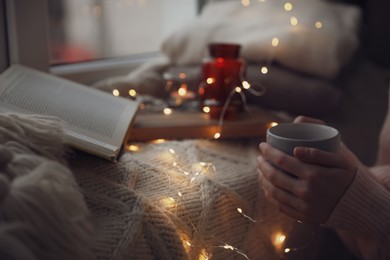 Photo of Woman with hot drink reading book near window at home, closeup. Cozy season