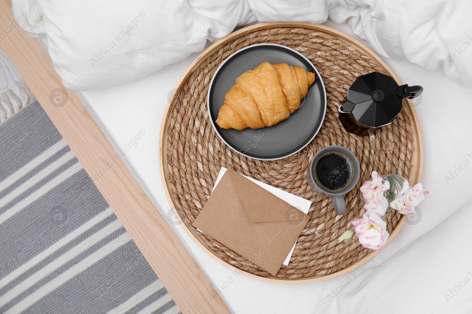 Photo of Tray with tasty croissant, cup of coffee and flowers on white bed, top view