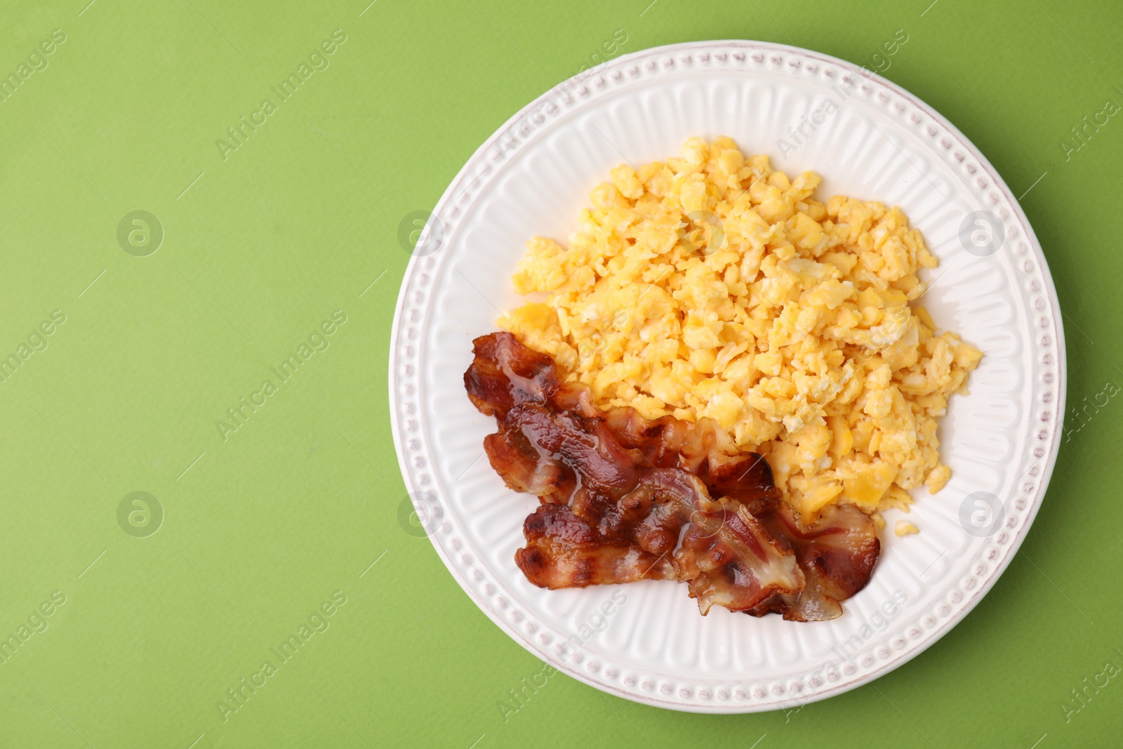 Photo of Delicious scrambled eggs with bacon in plate on light green background, top view. Space for text