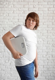Photo of Emotional overweight boy with floor scales near white brick wall