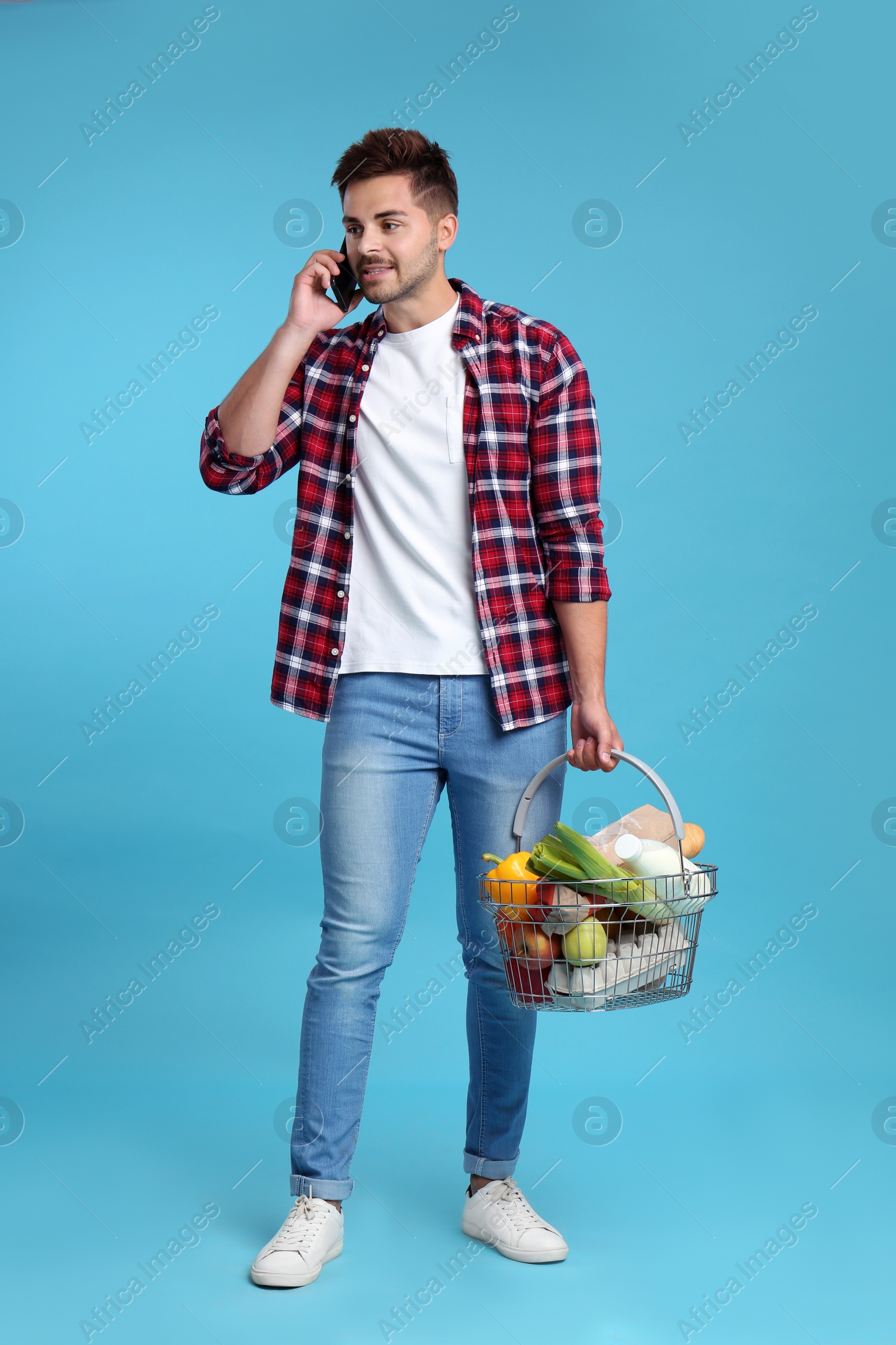 Photo of Young man holding shopping basket full of products while talking on phone against blue background