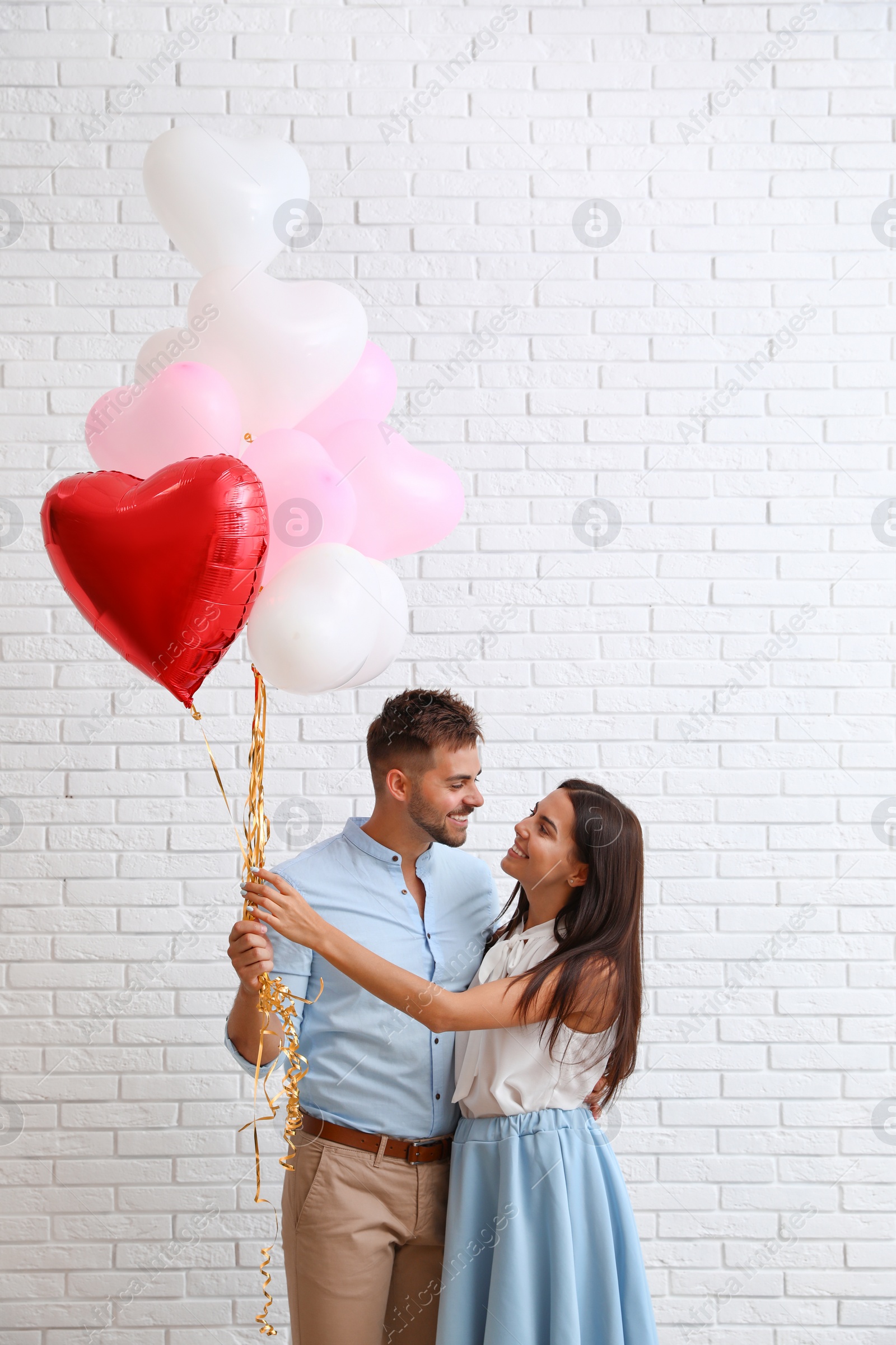 Photo of Young couple with air balloons near white brick wall. Celebration of Saint Valentine's Day