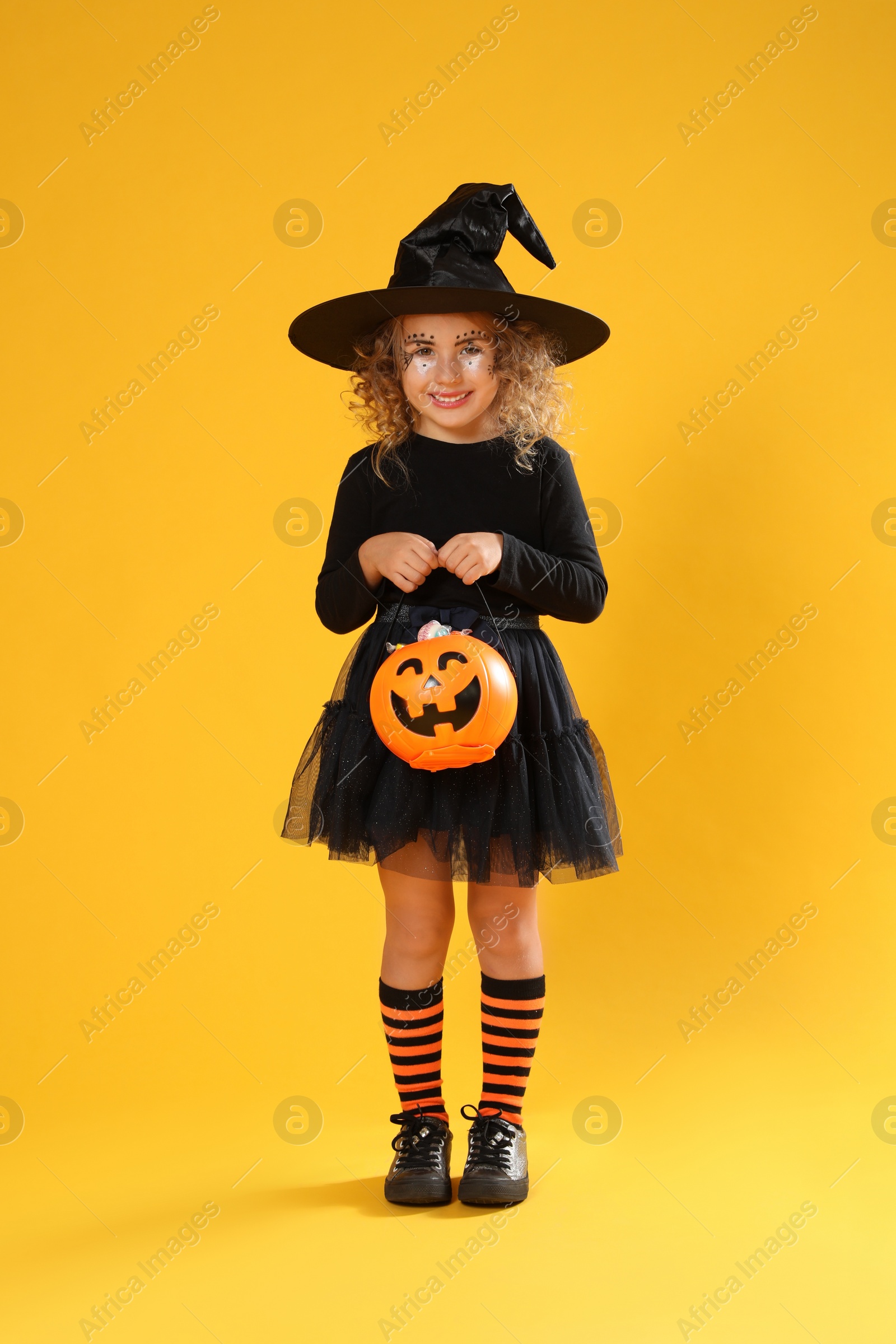 Photo of Cute little girl with pumpkin candy bucket wearing Halloween costume on yellow background