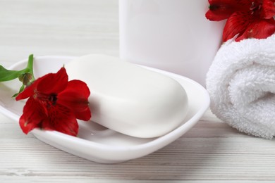 Photo of Dish with soap bar, terry towel and red flowers on white wooden table, closeup