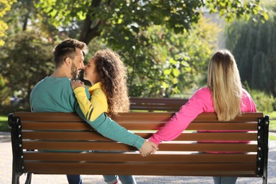 Photo of Man holding hands with another woman behind his girlfriend's back on bench in park. Love triangle