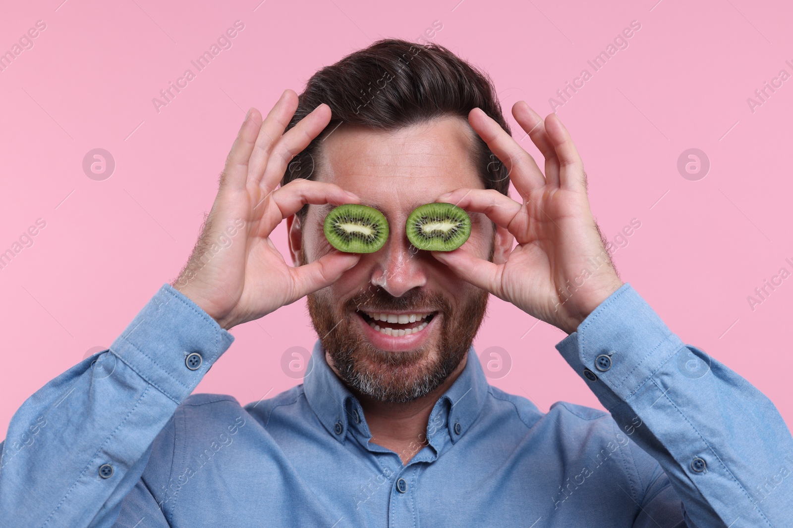 Photo of Man holding halves of kiwi near his eyes on pink background