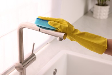 Photo of Woman in gloves cleaning faucet of kitchen sink with sponge, closeup