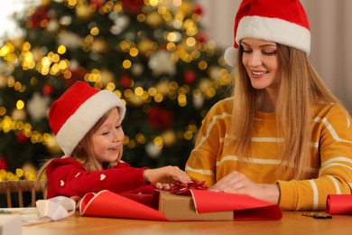 Photo of Mother and her little daughter in Santa hats wrapping Christmas gift at home