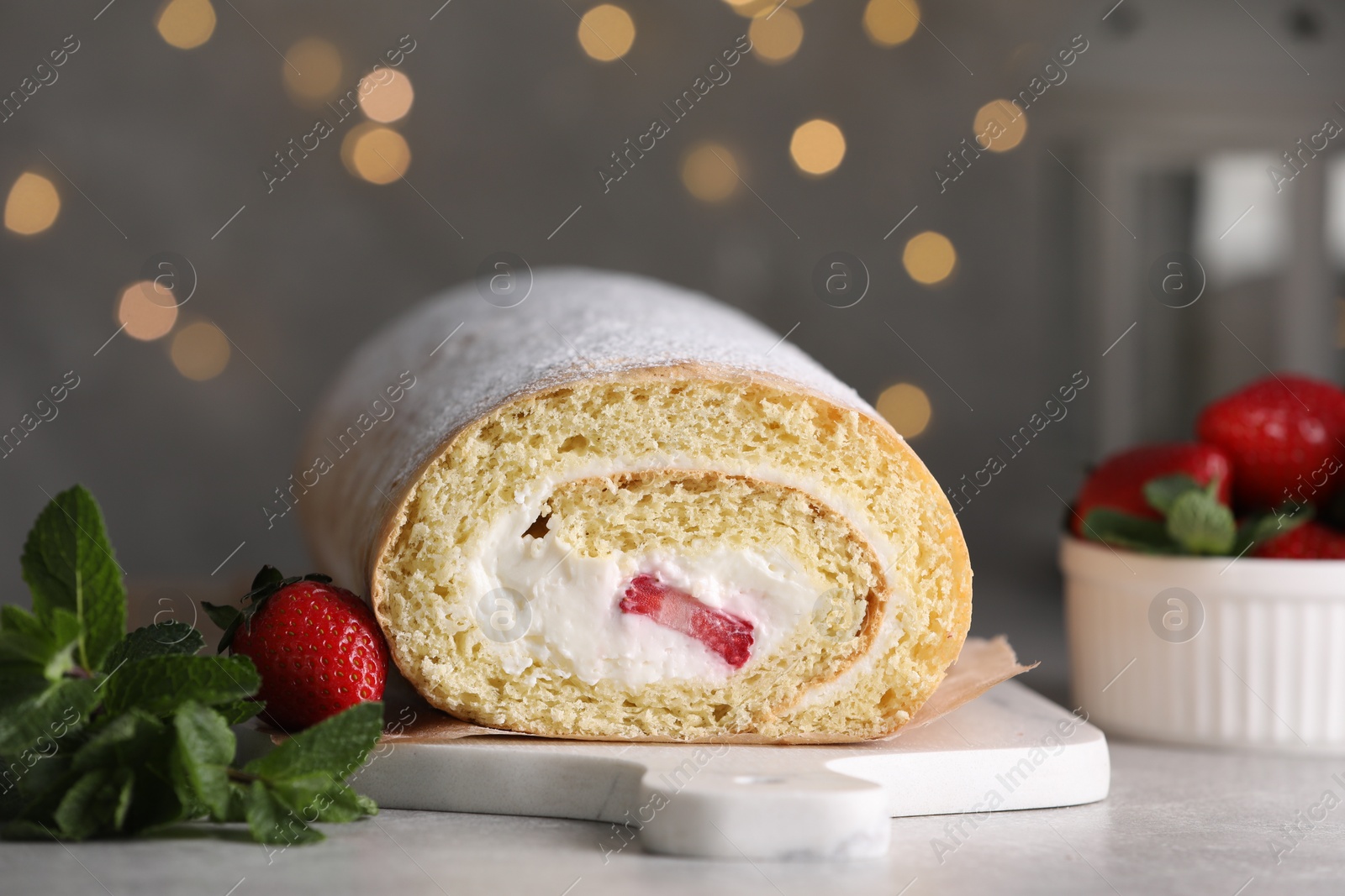 Photo of Delicious sponge cake roll with strawberries and cream on light grey table against blurred lights, closeup