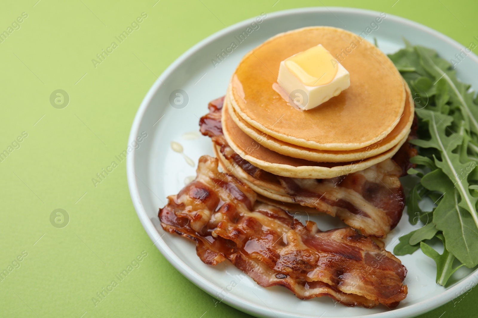 Photo of Tasty pancakes with butter, fried bacon and fresh arugula on light green background