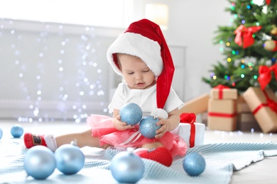 Cute baby in festive costume playing with Christmas decor on floor at home
