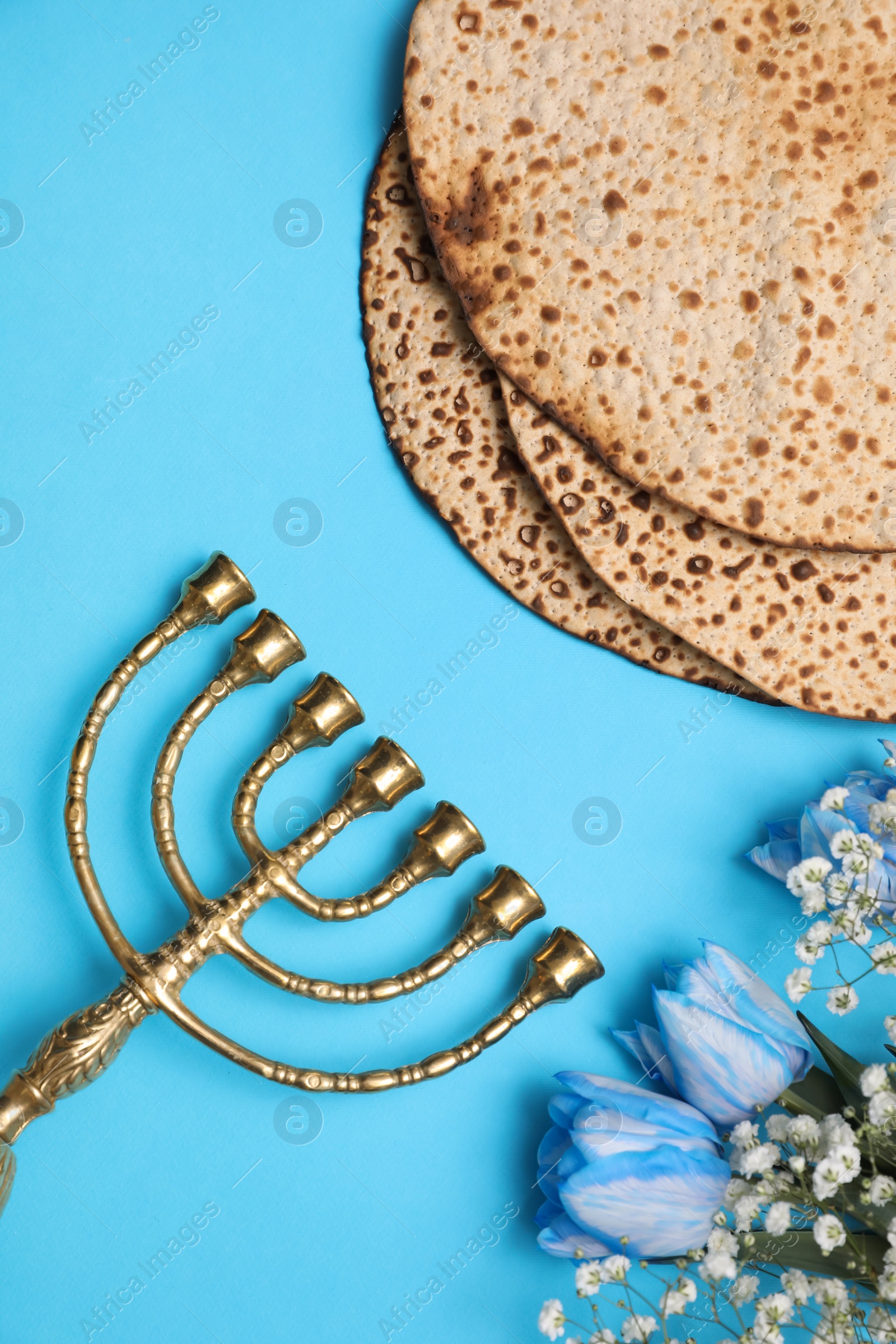 Photo of Tasty matzos, flowers and menorah on light blue background, flat lay. Passover (Pesach) celebration