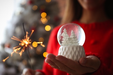 Woman with snow globe and sparkler near Christmas tree, closeup. Space for text