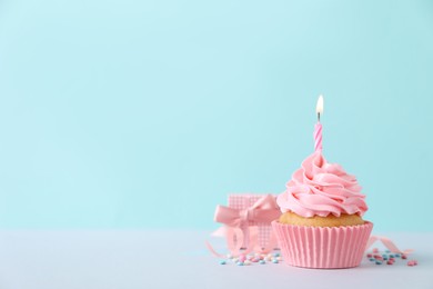 Delicious birthday cupcake with burning candle, sprinkles and gift box on white table against turquoise background, space for text