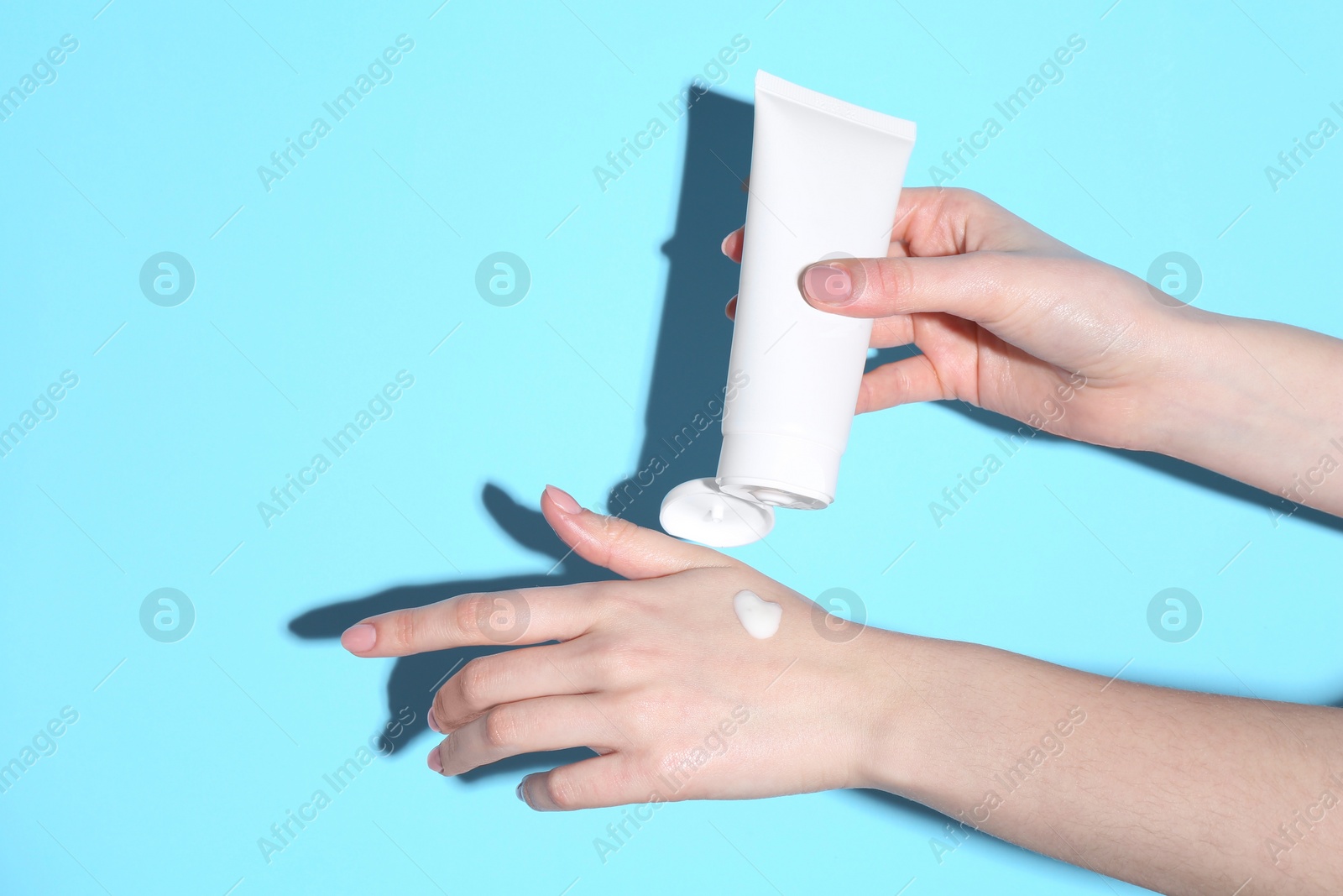 Photo of Woman with tube applying cream on her hand against light blue background, closeup