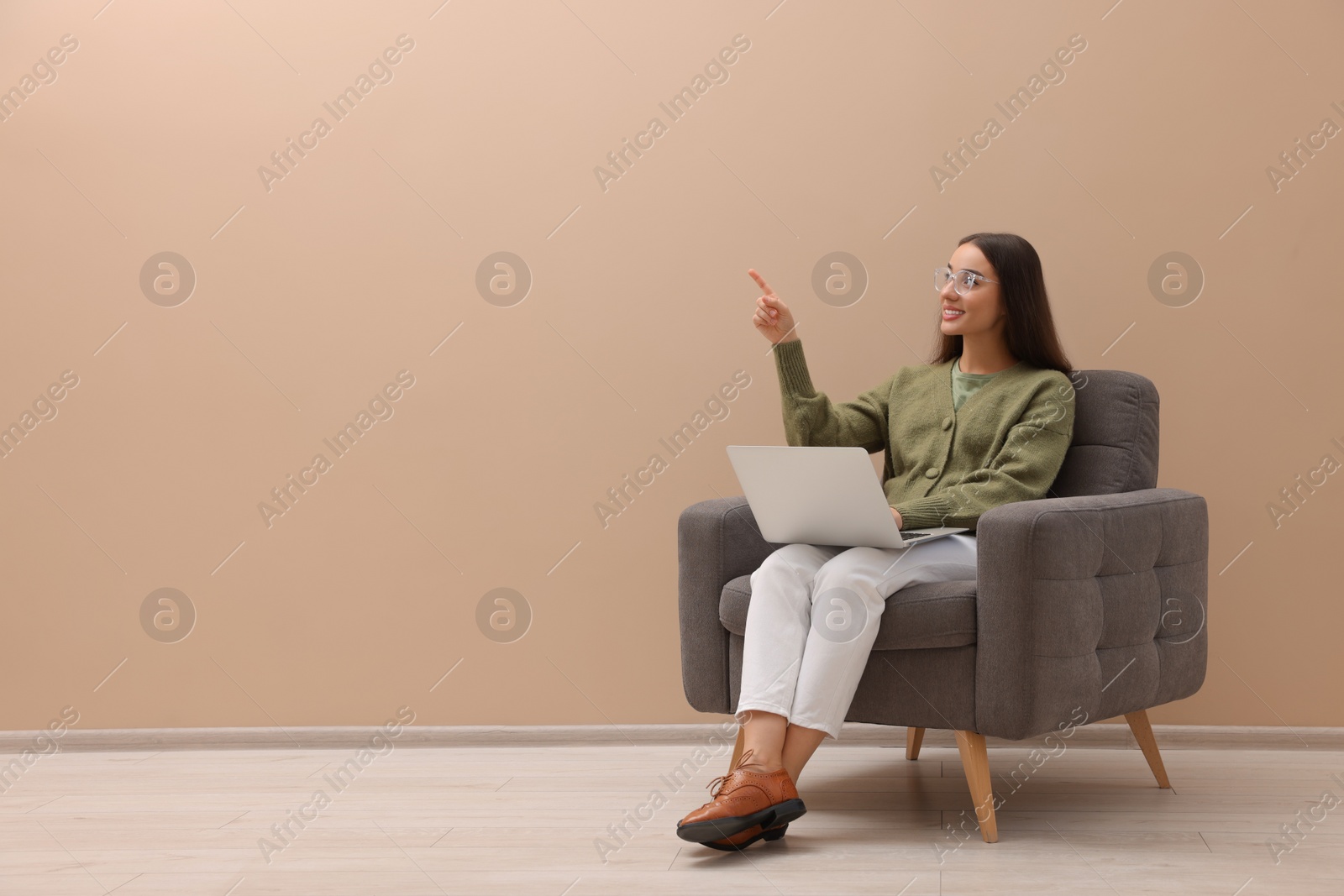 Photo of Beautiful woman with laptop pointing on something while sitting in armchair near beige wall indoors, space for text