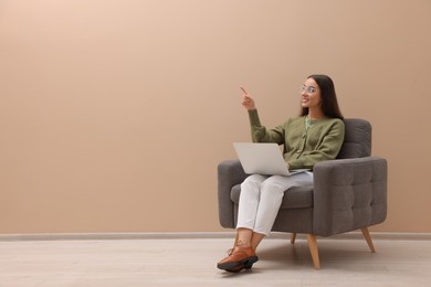 Beautiful woman with laptop pointing on something while sitting in armchair near beige wall indoors, space for text