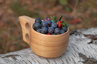 Wooden mug full of fresh ripe blueberries and lingonberries on log in forest, closeup